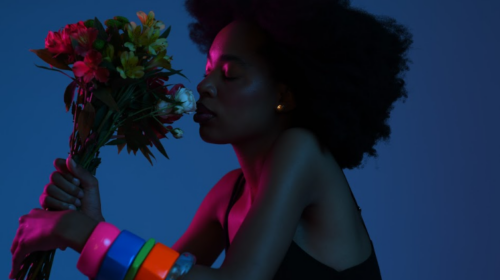 Foto de uma mulher negra de perfil. Ela está na frente de um fundo azul e está cheirando um buquê de flores. A mulher tem cabelo crespo e preto, usa várias pulseiras coloridas e está levemente iluminada por uma luz cor de rosa.