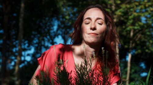 Imagem de uma mulher em uma área externa e arborizada. Ela é branca, em os cabelos ruivos e veste uma blusa cor de goiaba. Ela está cheirando flores de lavanda.