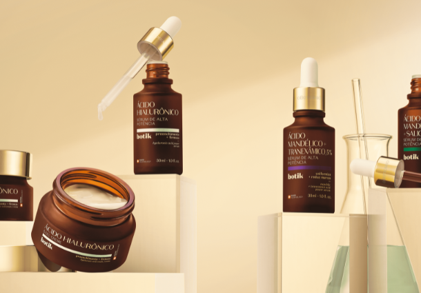 Quatro produtos da linha Botik de O Boticário são exibidos sobre uma superfície clara, com elementos de laboratório ao fundo. À esquerda, um pote de creme aberto e um pote fechado, ambos rotulados com "Ácido Hialurônico". Ao centro, um frasco âmbar com um conta-gotas aberto e outro frasco com um conta-gotas fechado, identificados como "Ácido Mandélico" e "Ácido Salicílico". Ao fundo, erlenmeyers com líquidos claros e esverdeados reforçam a atmosfera científica. A iluminação suave destaca os frascos âmbar e a textura dos cremes.
