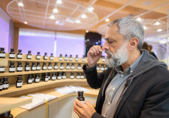 Um homem de perfil, com cabelos grisalhos e barba espessa, está absorto em sentir o aroma de uma tira de papel que segura perto do nariz. Em sua outra mão, ele segura um pequeno frasco âmbar. Ao fundo, prateleiras de madeira exibem inúmeros frascos idênticos, sugerindo um ambiente de perfumaria ou laboratório de aromas. A iluminação suave do local destaca a concentração do homem em sua avaliação olfativa. A cena transmite uma atmosfera de expertise e apreciação sensorial.