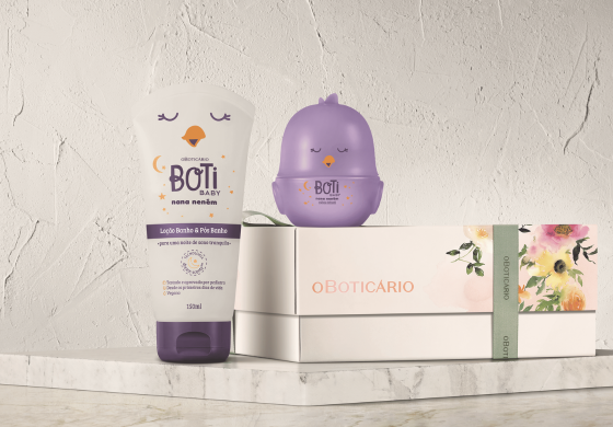Sobre uma superfície clara texturizada, um conjunto de produtos Boti Baby se apresenta. À esquerda, um tubo branco e roxo de loção com um desenho de lua crescente e estrelas. Ao centro, um recipiente roxo em formato de pintinho com olhos fechados. À direita, duas caixas brancas da O Boticário, uma delas adornada com uma faixa de papel floral. A composição sugere um presente delicado e voltado para cuidados infantis.