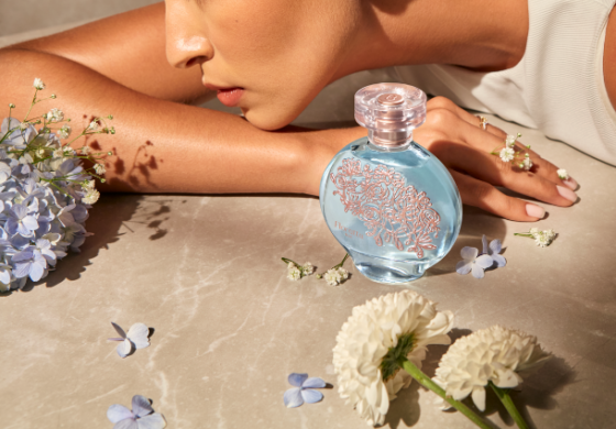 Perfume Floratta de O Boticário em primeiro plano na imagem, ao fundo o rosto de uma mulher apoiado sobre o seu braço, em volta há algumas flores e pétalas.