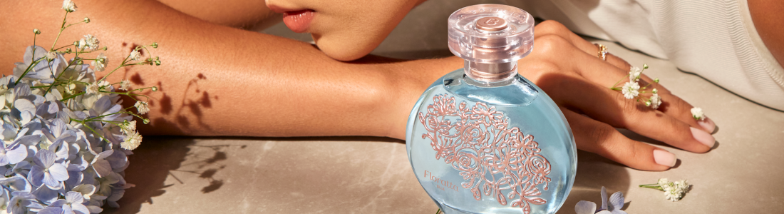 Perfume Floratta de O Boticário em primeiro plano na imagem, ao fundo o rosto de uma mulher apoiado sobre o seu braço, em volta há algumas flores e pétalas.