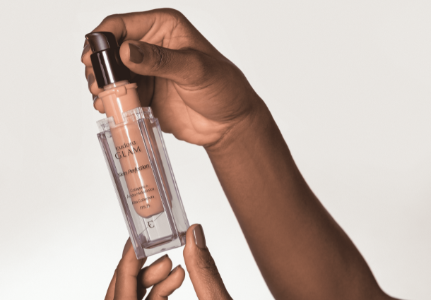 Duas mãos de pele morena seguram um frasco transparente de base líquida Glam de O Boticário contra um fundo branco suave. Uma mão apoia a base por baixo, enquanto a outra pressiona a bomba dispensadora preta. O líquido bege da base é visível através do frasco. A luz suave e difusa elimina sombras fortes, focando a atenção na embalagem elegante e na ação de usar o produto. A imagem transmite uma sensação de sofisticação e aplicação de maquiagem.