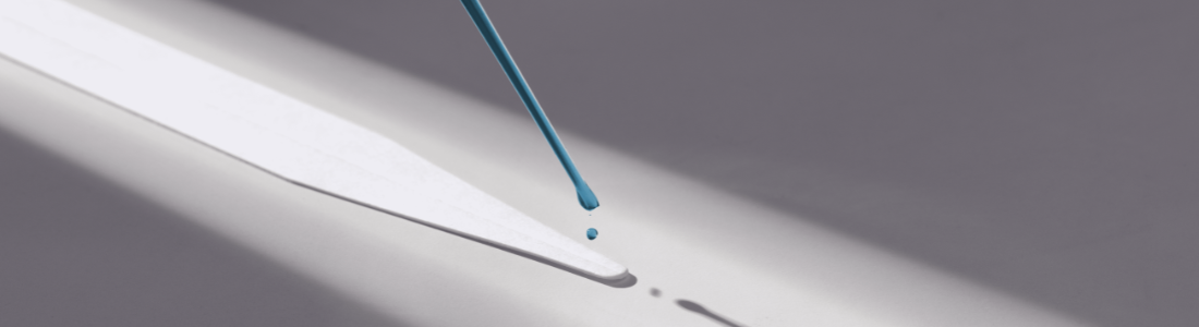Uma pipeta de vidro inclinada goteja um líquido azul intenso sobre uma superfície clara. Uma única gota alongada está prestes a cair, enquanto uma pequena poça já se formou abaixo. A luz lateral cria sombras marcadas da pipeta e da superfície. A cena minimalista destaca a precisão da ação em um ambiente de alto contraste. Evoca um momento de estudo ou experimentação cuidadosa.