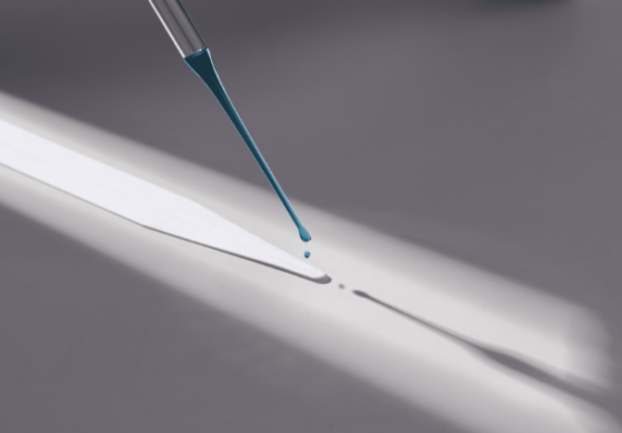 Uma pipeta de vidro inclinada goteja um líquido azul intenso sobre uma superfície clara. Uma única gota alongada está prestes a cair, enquanto uma pequena poça já se formou abaixo. A luz lateral cria sombras marcadas da pipeta e da superfície. A cena minimalista destaca a precisão da ação em um ambiente de alto contraste. Evoca um momento de estudo ou experimentação cuidadosa.