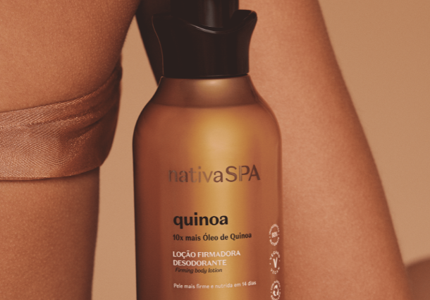 Um frasco dourado da loção firmadora desodorante Nativa SPA Quinoa de O Boticário se destaca contra a pele clara de uma pessoa. O frasco, com uma tampa preta, apresenta informações sobre os benefícios da quinoa e a quantidade de 400 ml. A luz suave incide sobre o frasco, realçando seu brilho metálico e a textura da pele ao fundo. A composição minimalista foca no produto e sua associação com o cuidado corporal. A imagem sugere nutrição e firmeza para a pele.