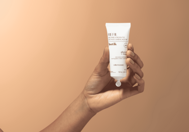 Uma mão de pele clara segura um tubo branco de creme facial Botik contra um fundo bege uniforme. O tubo apresenta informações em letras pequenas e um logotipo da marca O Boticário. A mão segura o tubo delicadamente, com o foco na embalagem do produto. A iluminação suave realça a textura da mão e a simplicidade do design da embalagem. A imagem transmite uma sensação de cuidado pessoal e minimalismo.