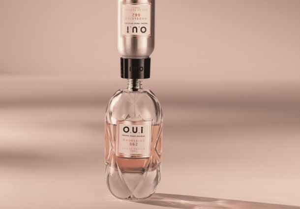Um frasco de perfume de vidro transparente, parcialmente preenchido com um líquido rosado, repousa sobre uma superfície bege. Acima dele, invertido e encaixado na abertura, encontra-se outro recipiente cilíndrico de metal prateado, também com detalhes rosados. Ambos exibem a inscrição vertical "O.U.i". A luz suave projeta uma sombra alongada do conjunto sobre a superfície. A composição minimalista destaca o design elegante e a conexão entre os dois elementos do produto.