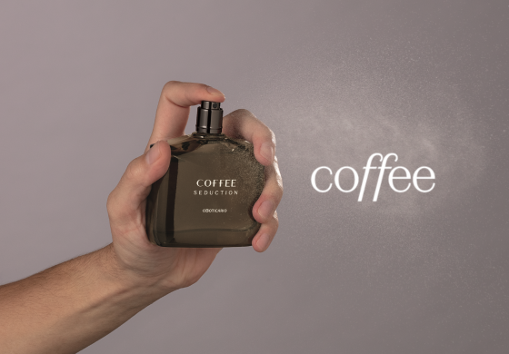 Mão borrifando o perfume Coffee Seduction, com a névoa visível sobre um fundo cinza suave.