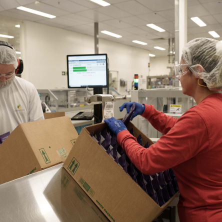 Na foto, há a imagem aproximada de duas mulheres trabalhando em uma unidade fabril, realizando o empacotamento das embalagens de produtos.