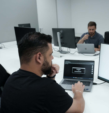 Na imagem, há a fotografia aproximada de um homem de pele branca, cabelos escuros e camiseta preta olhando concentrado para dois monitores, que mostram códigos de computação.