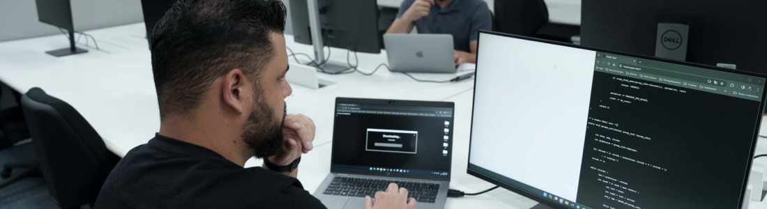 Na imagem, há a fotografia aproximada de um homem de pele branca, cabelos escuros e camiseta preta olhando concentrado para dois monitores, que mostram códigos de computação.