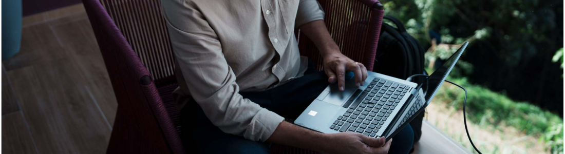 Na foto, há a imagem aproximada das mãos de um homem com pele negra, camisa social azul e calça social preta utilizando um notebook.