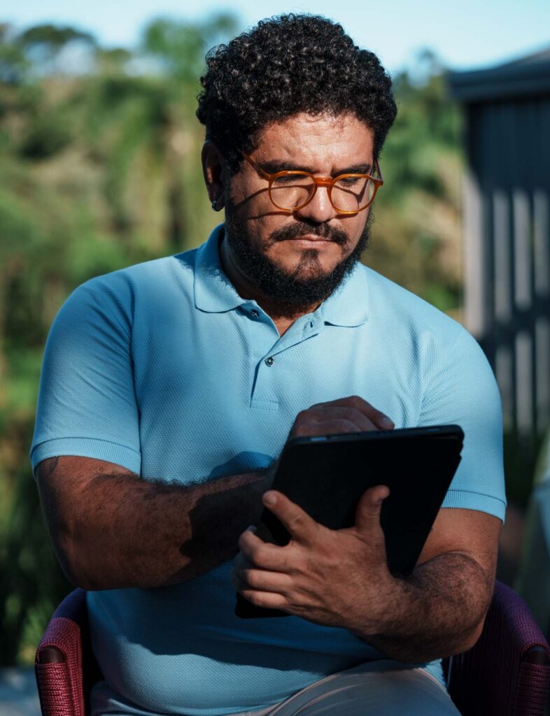 Na foto, há a imagem aproximada de um homem de meia idade, pele branca, camisa polo azul e óculos com armação na cor laranja. Com semblante concentrado, ele olha para a tela de um tablet.