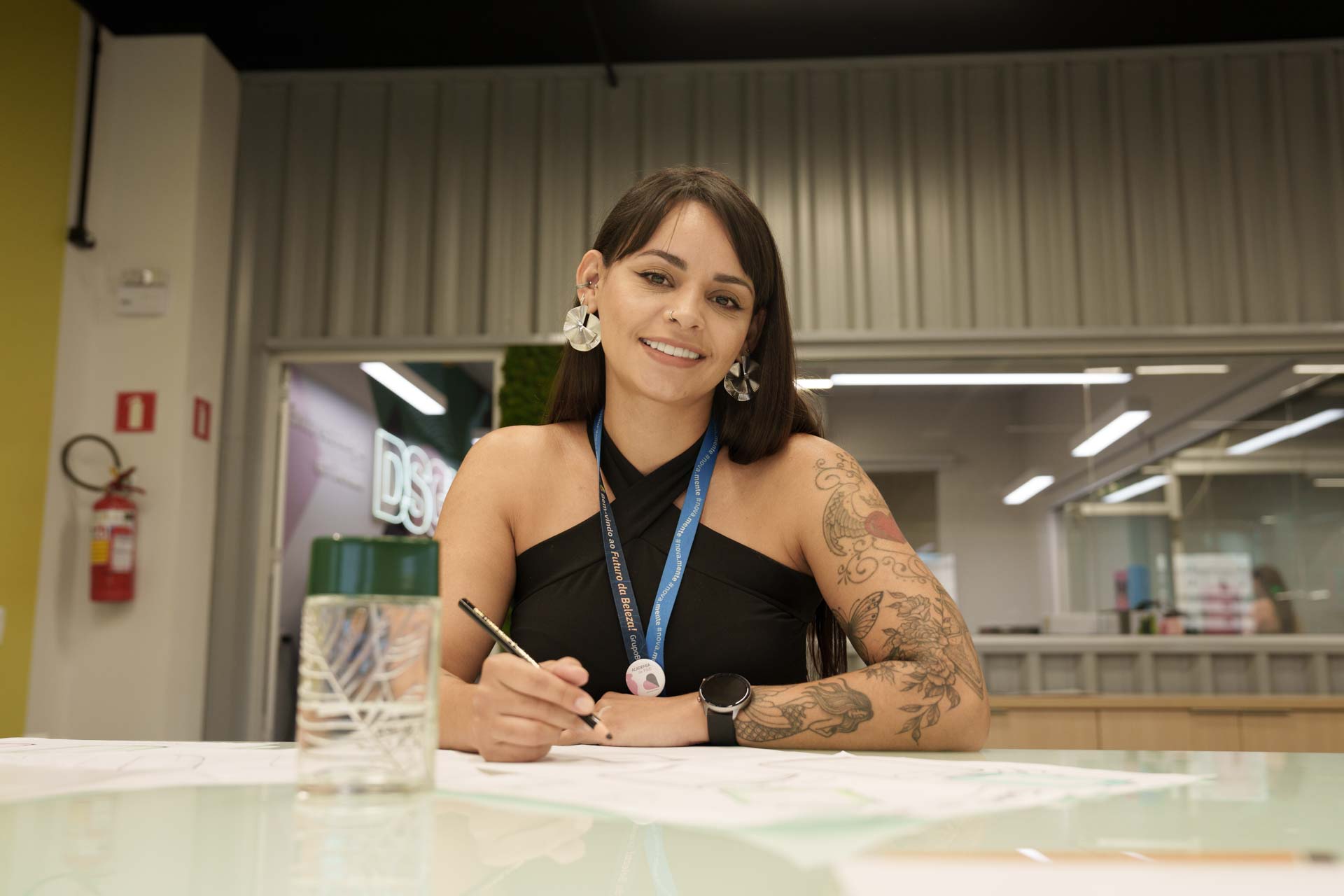 Na foto, há a imagem de uma mulher de pele branca com tatuagens, cabelos escuros e roupa preta. Seus braços estão apoiados em uma mesa de escritório, com um lápis na mão esquerda. Ela sorri para a câmera.
