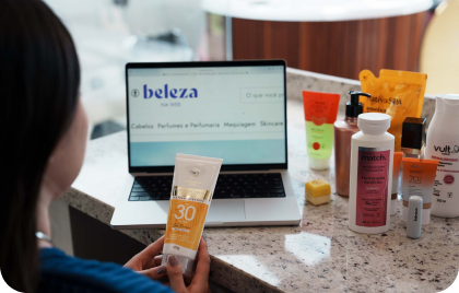 Na foto, há a imagem aproximada de uma mulher analisando o rótulo de um dermocosmético e, ao fundo, há um notebook com a página aberta do site Beleza na Web.

