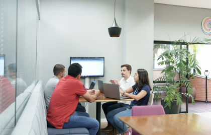 Na foto, há a imagem aproximada de um grupo de pessoas reunidas em uma mesa de escritório, sendo três homens e uma mulher.
