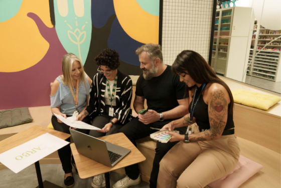 Na foto, há a imagem aproximada de um grupo de pessoas com diferentes idades, sendo três mulheres e um homem. Todos estão sentados em um banco de madeira localizado em uma área externa, conversando animadamente.