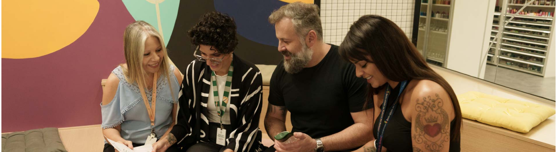 Na foto, há a imagem aproximada de um grupo de pessoas com diferentes idades, sendo três mulheres e um homem. Todos estão sentados em um banco de madeira localizado em uma área externa, conversando animadamente.