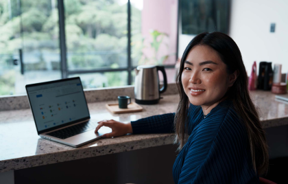 Na foto, há a imagem aproximada de uma mulher com traços asiáticos, pele branca e cabelos escuros sorrindo para a câmera. Ao fundo, há uma mesa de escritório com um notebook sobre ela.
