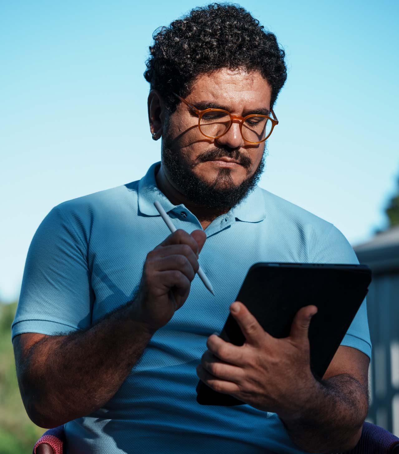 Na foto, há a imagem aproximada de um homem de meia idade, pele branca, camisa polo azul e óculos com armação na cor laranja. Com semblante concentrado, ele olha para a tela de um tablet.
