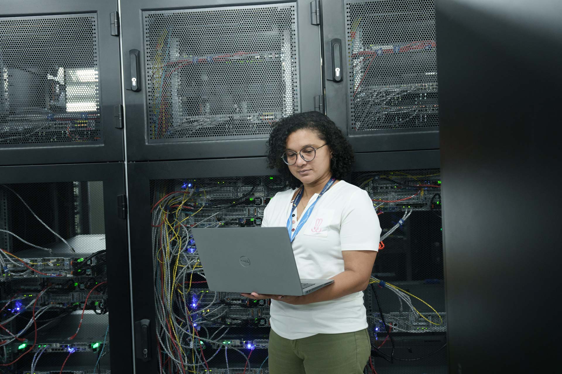 Na foto, há a imagem de uma mulher de pele negra, cabelos escuros, camisa branca e calça verde em pé, enquanto segura um notebook. Ela olha atentamente para a tela.
