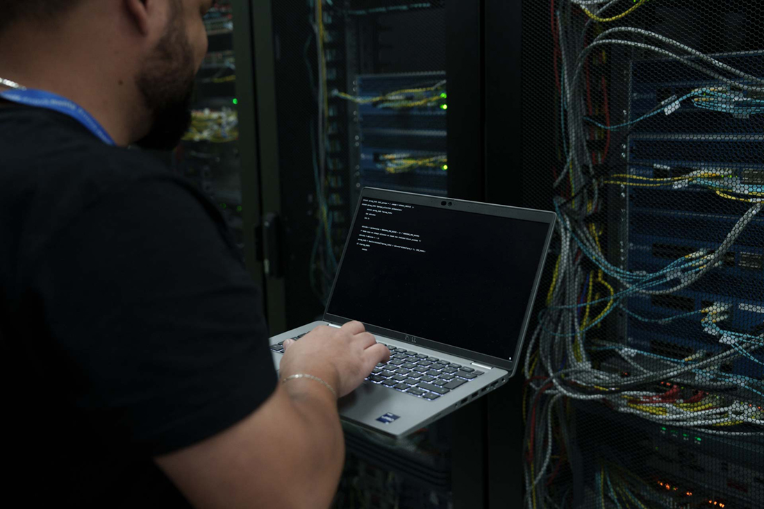 Na foto, há a fotografia aproximada de um par de mãos mexendo em um notebook. Ao fundo, há luzes e fios que lembram uma estação de computadores.