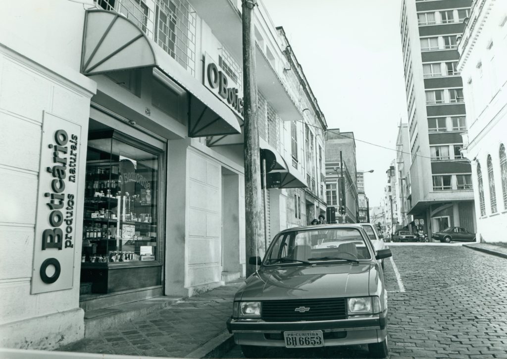 Imagem em preto e branco da frente da antiga farmácia de manipulação. Na faixada da loja está descrito O Boticário Produtos Naturais. Em frente a loja tem um carro 