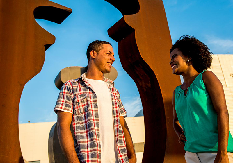 Foto de um homem e uma mulher em frente a fabrica de Camaçari, na Bahia. Eles estão conversando. Ele, veste uma camiseta branca com uma camisa xadrez por cima. Ela veste uma regata azul. Atrás deles tem a ânfora de O Botícario