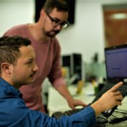 Homens olhando e mexendo nos computadores. Na frente, homem de azul com barba e cabelos castanhos escuros. Atrás, homem de camisa rosa, ele usa óculos e também tem barba e cabelos castanhos escuros