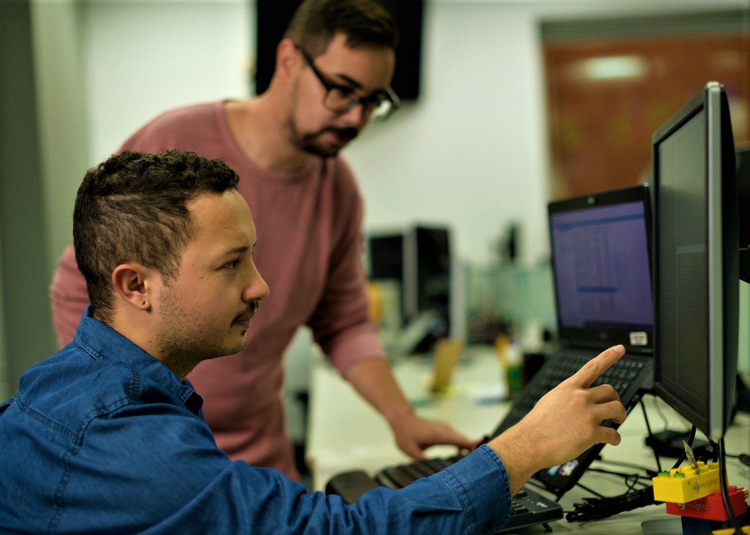 Homens olhando e mexendo nos computadores. Na frente, homem de azul com barba e cabelos castanhos escuros. Atrás, homem de camisa rosa, ele usa óculos e também tem barba e cabelos castanhos escuros