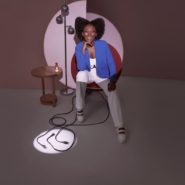 mulher negra sorrindo ao centro da imagem, sentada em uma cadeira circular, vestida de camisa, calça e tênis branco e um blaizer azul escuro. A imagem destaca somente a mulher e os fios soltos, o restante está em sombra