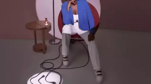 mulher negra sorrindo ao centro da imagem, sentada em uma cadeira circular, vestida de camisa, calça e tênis branco e um blaizer azul escuro. A imagem destaca somente a mulher e os fios soltos, o restante está em sombra