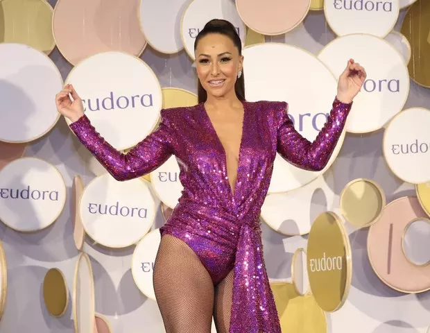 Foto da Sabrina Sato em frente ao fundo com balões de Eudora. Ela veste um maiô de manga longa e rosa, com cabelos castanhos longos e preso com rabo de cavalo