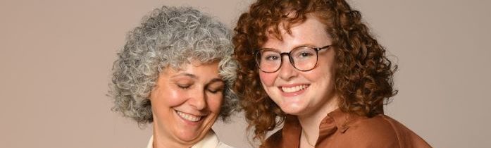 Imagem de duas mulheres sorrindo. Da esquerda para direita temos uma mulher, aparenta ter 40+, com cabelos grisalhos e está olhando para baixo. Ao seu lado uma mulher usando óculos, cabelos em tons avermelhados e usa camisa marrom