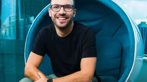 Imagem de Daniel Knopfholz. Homem, branco, usa óculos e camiseta preta. Esta sentado em uma cadeira azul redonda, ele sorri para foto