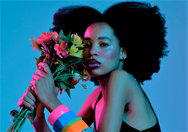 Mulher negra na frente de um fundo azul. Ela tem o cabelo afro e veste regata preta. Esta segurando um buquê de flores colorido.