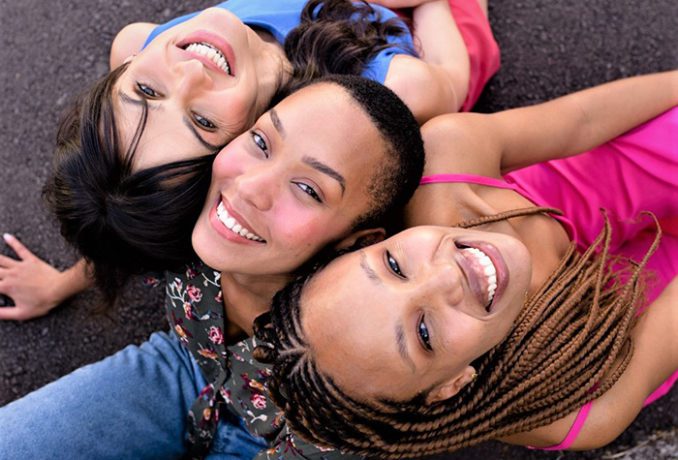 Imagem com três mulheres sorrindo e olhando para cima. Da esquerda para direita temos uma mulher branca vestindo camiseta azul, ao lado dela temos uma mulher negra careca, ela usa uma camisa florida. Ao lado direito temos uma mulher negra com trancas cor caramelo, ela veste uma camiseta regata rosa