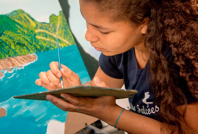 Imagem de mulher negra pintando quadro. Atrás dela tem um quadro de paisagem, ela está com um pincel e uma tela na mão