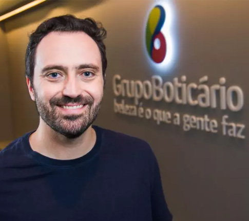 Imagem de José Ronchetti. Homem, branco, com barbas e cabelos castanhos escuros. Ele sorri para imagem e está em um ambiente corporativo.