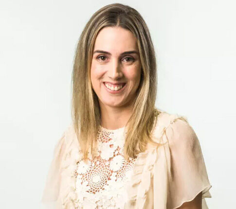 Imagem Renata Gomide, Mulher branca, loira, veste camisa cor-de-rosa bem clarinho e está sentada em uma cadeira alta sorrindo