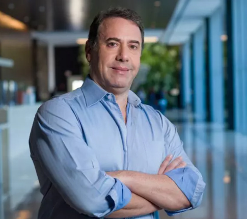 Imagem de Artur.Homem, branco, cabelos escuros. Ele veste camisa azul, está de braços cruzados em ambiente corporativo.