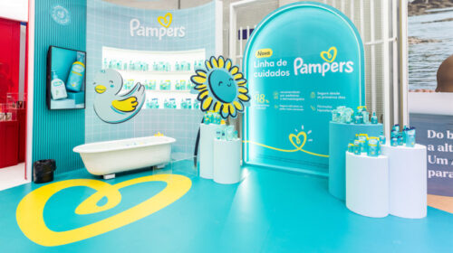 Imagem do stand de Pampers, com a exposição da nova linha que possui 98% de ingredientes naturais. A imagem tem uma estrutura de banheiro de bebê azul, com patinhos e flores nas cores da marca, além dos produtos de lançamento espalhados no stand.