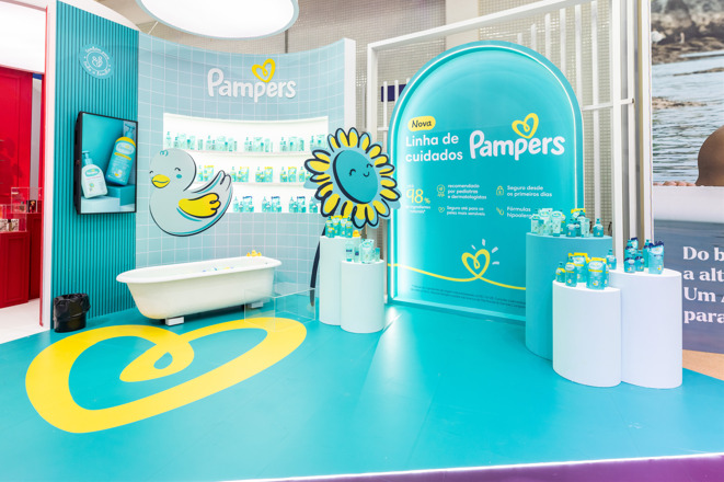Imagem do stand de Pampers, com a exposição da nova linha que possui 98% de ingredientes naturais. A imagem tem uma estrutura de banheiro de bebê azul, com patinhos e flores nas cores da marca, além dos produtos de lançamento espalhados no stand.