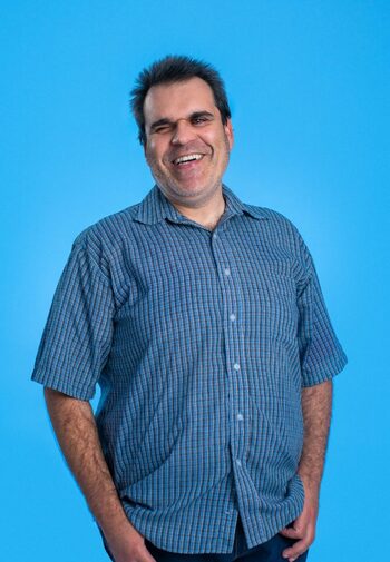 Image of a white man with visual impairment. He is in front of a blue background and wears a plaid shirt also in blue tones.