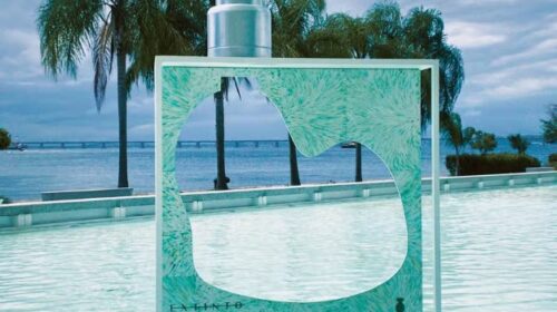 Imagem de um frasco de perfume gigante de O Boticário, do Projeto Extinto, para conscientização ambiental. O frasco está posicionado sobre um espelho d’água. O frasco é verde água e a válvula spray prateada. No centro do frasco há um recorte vazado no formato da Baía de Guanabara. Ao fundo, uma paisagem de céu azul, mar e coqueiros.