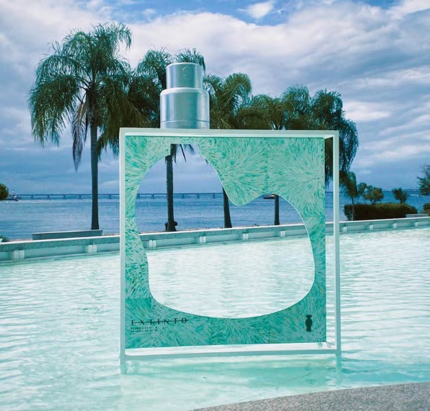 Imagem de um frasco de perfume gigante de O Boticário, do Projeto Extinto, para conscientização ambiental. O frasco está posicionado sobre um espelho d’água. O frasco é verde água e a válvula spray prateada. No centro do frasco há um recorte vazado no formato da Baía de Guanabara. Ao fundo, uma paisagem de céu azul, mar e coqueiros.
