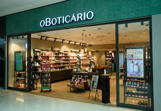 Foto da fachada de uma loja de O Boticário. A testeira da loja é verde e “O Boticário” está escrito em branco. Há uma vitrine de cada lado da loja, com posters em destaque e produtos em exposição. Dentro da loja, há várias estantes com produtos de embalagens de várias cores diferentes.