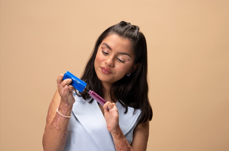 Imagem de uma mulher em fundo laranja claro. Ela é branca, tem cabelos lisos e castanhos na altura dos ombros, usa maquiagem colorida e uma blusa branca. A mulher é uma pessoa com deficiência nas mãos e está segurando um dos acessórios inclusivos do Grupo Boticário, o engrossador multiuso, na mão direita e na mão esquerda está uma máscara de cílios.